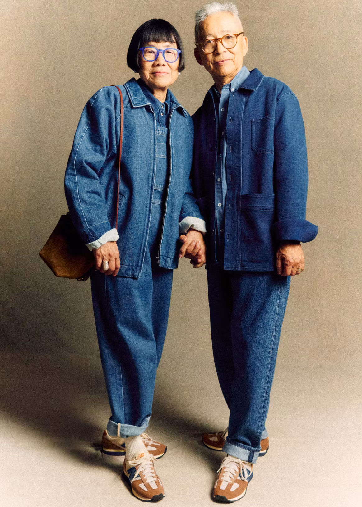 A full length photo of a male and female model holding hands. They both wear top to toe denim and glasses, and the woman is holding a brown bag. Their jeans are rolled up revealing the sneaker collaboration design