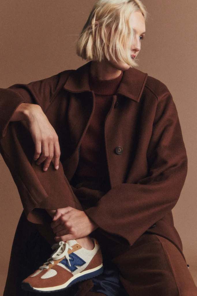 A blonde female model wears the New Balance Sézane 471 design. She is dressed all in brown, and sat with her leg bent up to reveal the sneaker