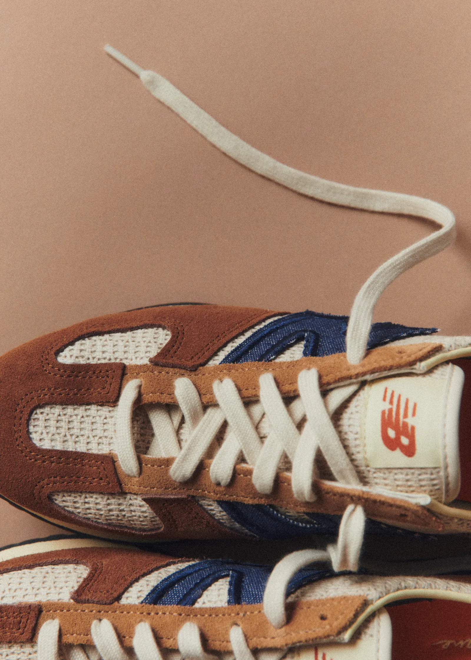 The New Balance Sézane 471 sneaker design viewed from above against a mocha coloured background. The sneakers shoelaces are untied and draped loosely down the sides