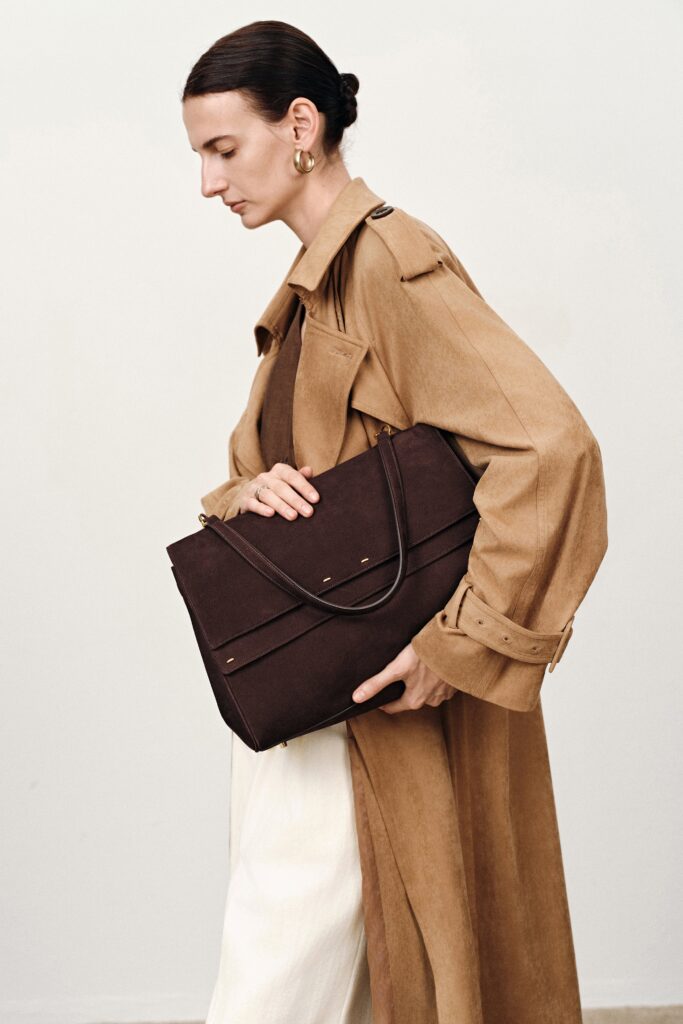 A woman in a camel mac looking down carrying a quiet luxury bag by fabrique under her arm. The bag is brown suede and large. Her hair is tied up and she's also wearing white jeans. She's photographed against a cream background.