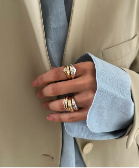 Close up photo of a woman's hand wearing multiple stacked mix metal jewellery rings. You can see the cuff of her blue shirt and a stone coloured blazer